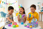 Three children laughing and playing with colorful squishy toys on a white table in a bright, decorated room. A blue retail display box for the toys is visible on the table.