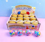 A display box filled with small round toy buns labeled 'Mysterysquishy Steameo Bun,' with four colorful round squishy balls in front. The toys are photographed on a pastel pink surface against a light blue starry backdrop.