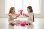 Two young girls sitting on a wooden floor in a bright room, playing with a pink toy hair dryer and makeup accessories. One girl holds a pink hair dryer, and the other holds a pink makeup brush, with a pink toy makeup bag and scattered makeup pieces in front of them.