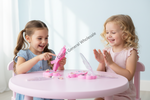 Two young girls sitting at a pink round table, smiling and playing with a pink butterfly-themed craft or activity set. The scene is brightly lit with soft window light, and a diagonal watermark reading 'Dallas General Wholesale' is visible across the center.