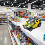 A toy remote control sports car set displayed on a toy store shelf, featuring a yellow and green remote-controlled off-road vehicle, a black remote controller, and its retail packaging box. The background shows a busy toy store aisle with shelves of other toys.