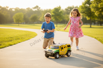 A young boy in a blue shirt and khaki pants is kneeling down, smiling excitedly while holding a remote control. Next to him, a young girl in a pink floral dress is running and laughing on a paved path in a sunny park.