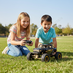A young girl and boy are lying on a green grass field, smiling as they play with a black and gold remote control off-road truck. The girl holds a black remote control, and the boy looks at the truck.