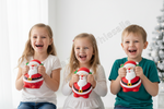 Three children sitting side by side, each holding a small Santa Claus-shaped plush toy. The children are smiling and laughing in a bright indoor setting with a Christmas tree visible in the background.
