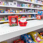 A red Santa Claus squishy toy next to its original red packaging box on a white toy store shelf, with a blurred background of colorful toys.