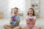 Two children sitting cross-legged on a rug in a bright living room, each holding a purple textured stress ball and laughing. A watermark for Dallas General Wholesale is repeated across the image.
