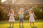 Three children playing outdoors on a grassy lawn with bubbles in the air. The child on the left wears a colorful striped dress, the center child holds a yellow bubble blower, and the child on the right wears a white floral dress.