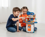 Two children sitting on a light floor playing with a colorful toy play kitchen set in a bright room.