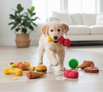 A golden retriever puppy stands on a light wood floor in a bright living room, holding a red and yellow toy boxing glove in its mouth. Several colorful dog toys are scattered around the floor, including a yellow bone, a hamburger toy, a green spiky ball, and a brown chicken leg toy.
