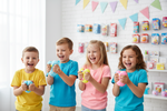 Four children laughing and playing with colorful slime in a bright, decorated toy store or playroom. The children are wearing yellow, blue, pink, and teal t-shirts, holding light blue, yellow, and pink slime.
