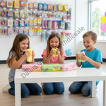 Three children sitting on the floor around a white table, each holding a colorful squishy toy while looking at their branded boxes on the table. The background shows a toy store interior with shelves of products.