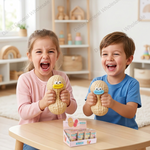 Two children, a girl in a pink cardigan and a boy in a blue t-shirt, laughing while squeezing peanut-shaped toys with colorful beaks. A small display box of similar toys sits on the table in front of them, set in a bright, modern children's room.