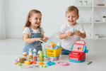 Two young children playing with a colorful toy grocery store set on a white floor. The set includes a yellow shopping cart, a red and blue toy cash register, small pretend bottles, produce, and play money.