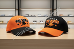 Two baseball caps displayed side by side on a light wood counter in a store setting. The left cap is orange and black with a black brim and black embroidered 'TEXAS' text with a cowboy silhouette.