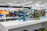 Two toy electric blasters displayed on a store shelf, one blue with orange accents and one green with orange accents, with their retail packaging behind them and a flame effect edited onto the left blaster.