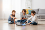 Three children sitting on a wooden floor in a bright living room, playing with a silver remote-controlled toy car with blue LED lights. One child holds a black controller.