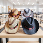 Four cowboy-style hats displayed on a wooden counter in a store interior. The hats are in black, tan, cream, and gray, each featuring a red floral band with a silver-toned cross emblem.