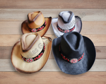 Four cowboy hats arranged on a light wood plank background. The hats are shown in multiple colors: brown, tan, white, and black, each featuring a black band with red floral embroidery and a silver-toned circular buckle.