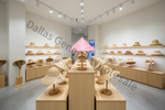 A retail display of straw and woven wide-brim hats arranged on wooden pedestals and wall shelves in a bright, modern store interior. A pink patterned awning-like shade sits on a central pedestal, and multiple hat styles are visible.